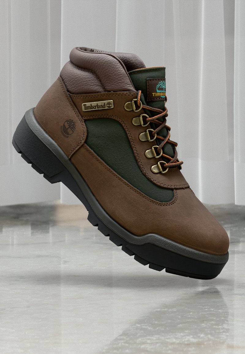 Botte de randonnée Timberland marron et verte avec col rembourré, œillets métalliques, lacets et semelle noire robuste sur une surface en marbre.