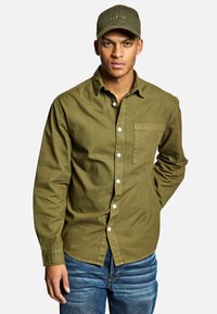 Camicia button-up verde oliva con tasca sul petto, abbinata a jeans blu. La camicia ha un colletto classico e polsini con bottoni. Viene indossato un cappellino coordinato.