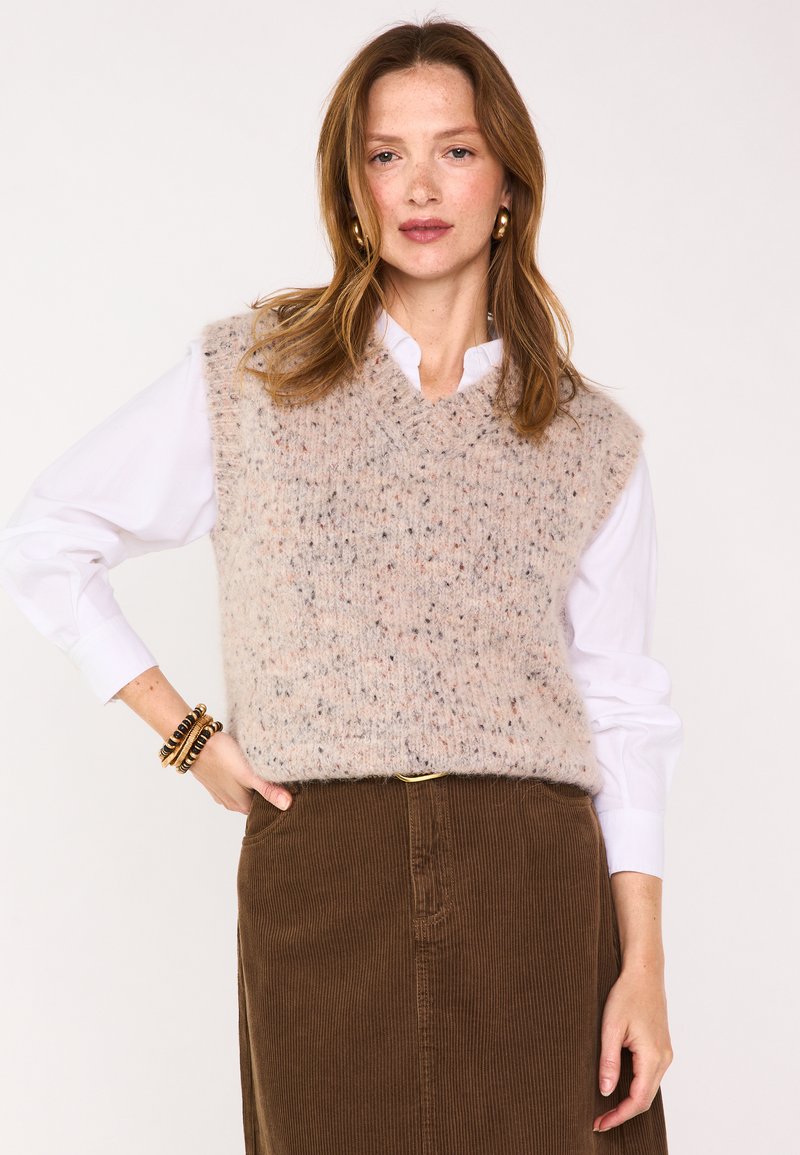Gilet en maille à col en V en laine douce beige clair avec des touches de couleur. Porté sur une chemise blanche et associé à une jupe en velours côtelé marron.