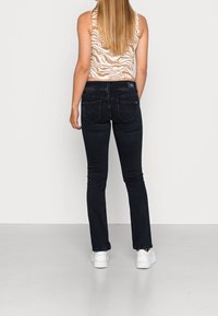 Kvinna med långt hår, iklädd en ärmlös topp med brun och vit zebramönster, mörka skinny jeans och vita sneakers, stående mot en enkel vägg.