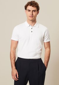 Camisa polo blanca de manga corta con collar clásico, tres botones negros y un diseño ajustado, combinada con pantalones navy.