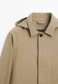 Beige gewatteerde jas met capuchon, puntige kraag en één zichtbare zwarte knoop aan de voorkant, afgebeeld tegen een witte achtergrond.