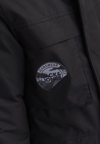Svart jacka med en rund, upphöjd patch med texten "Jack & Jones". Materialet är slätt med en matt yta och synlig sömnad.