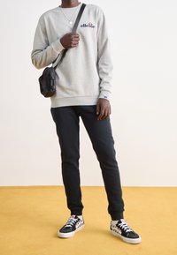 Sweat-shirt gris avec logo bleu marine, associé à un pantalon de survêtement noir cintré. Chaussures à motif damier noir et blanc. Sac bandoulière noir.