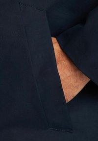 Hand delvis inskjuten i fickan på en mörk marinblå kavajärm.