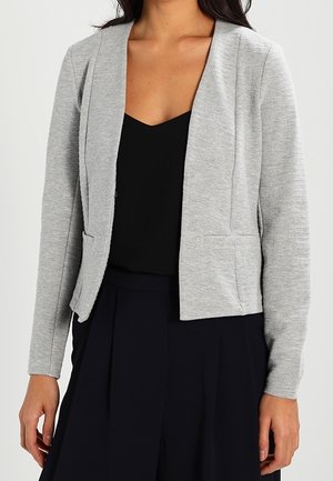 Kvinde iført en lysegrå åben-front blazer over en sort V-hals top, kombineret med højttaljede sorte plisserede bukser.