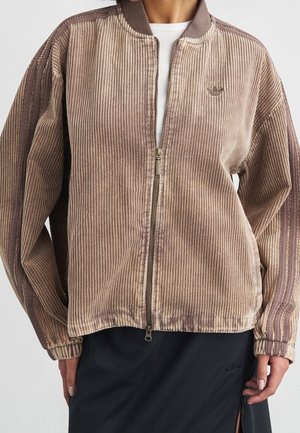 Fløjlsjakke med lynlås i beige og brune nuancer båret over en hvid top, kombineret med et sort skørt med en sidesplit.