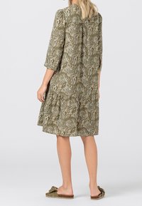 Nainen, jolla on päällään polvenpituinen oliivinvihreä paisley-mekko, jossa on kolmasosa-hihattomat hihat, sekä oliivinvihreät liukuvat sandaalit, joissa on koristeellinen nauha.