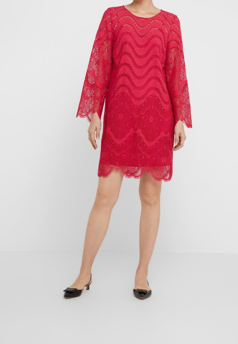 Femme portant une robe rouge en dentelle longueur genou avec des bords festonnés et des manches trois-quarts, assortie à des chaussures noires à talons bas.