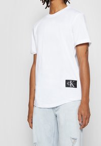 Biała bawełniana koszulka z krótkim rękawem, okrągłym dekoltem i czarną naszywką logo Calvin Klein na lewej biodrze. Jasne dżinsy o zniszczonym wyglądzie.