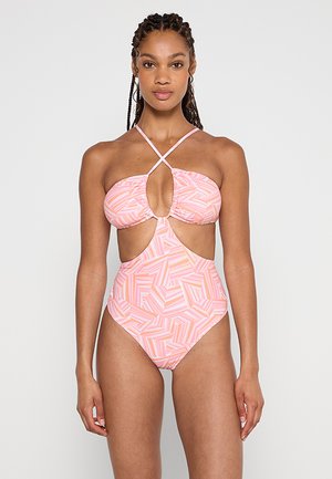 Woman with braided hair wears pink and orange geometric patterned one-piece swimsuit with front cutouts and crisscross halter straps.