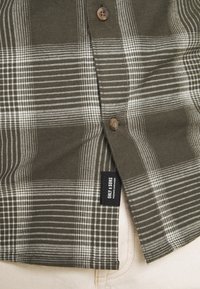 Grün-weiß kariertes Hemd mit Knopfleiste, braunen Knöpfen und einem schwarzen Label nahe dem Saum. Hergestellt aus einem strukturierten Stoff.