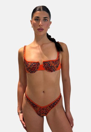 Donna con capelli scuri intrecciati che indossa un bikini arancione e nero a motivo, in piedi davanti a uno sfondo chiaro uniforme, guarda dritto davanti a sé.