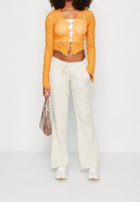 Femme portant un cardigan court en dentelle orange avec un débardeur blanc en dessous, un pantalon beige à cordon de serrage, des baskets blanches, et tenant un sac à main en cuir de serpent.