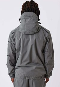 Giacca cappuccio grigia con una texture liscia, polsini elasticizzati e cappuccio con cordino. Presenta dettagli neri e una tasca sul petto con zip.