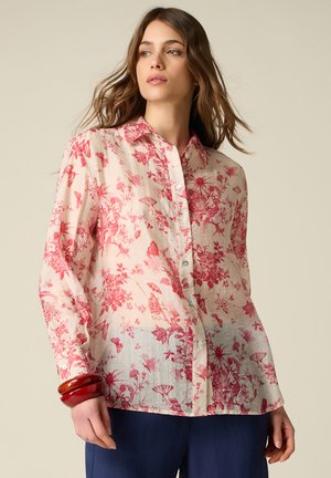 Vrouw draagt een lichtroze blouse met knopen en rode bloemen- en vlinderprint, gecombineerd met donkerblauwe broek en rode armbanden om de pols.