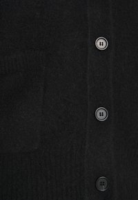 Černý kardigan z vlny s měkkou texturou, s kapsou a šesti černými knoflíky podél přední části. Lemování s žebrováním na okrajích.