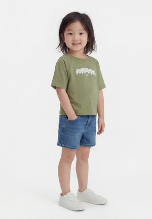 Μικρό παιδί που στέκεται με τα χέρια στις τσέπες, φορώντας πράσινο μπλουζάκι με λουλούδια, τζιν σορτς και άσπρα αθλητικά παπούτσια, μπροστά σε απλό λευκό φόντο.