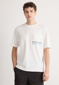 Bílý bavlněný tričko s přední kapsou a barevným logem "HUGO". Krátké rukávy, uvolněný střih a hladká textura.