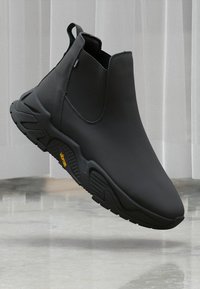Black ankle boot with a sleek design, featuring a textured sole, displayed against a light backdrop with soft fabric draping in the background.