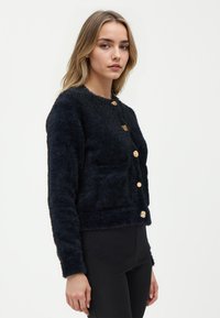 Cardigan nero e accogliente realizzato in un morbido tessuto testurizzato con bottoni dorati e scollatura rotonda, caratterizzato da una silhouette aderente.