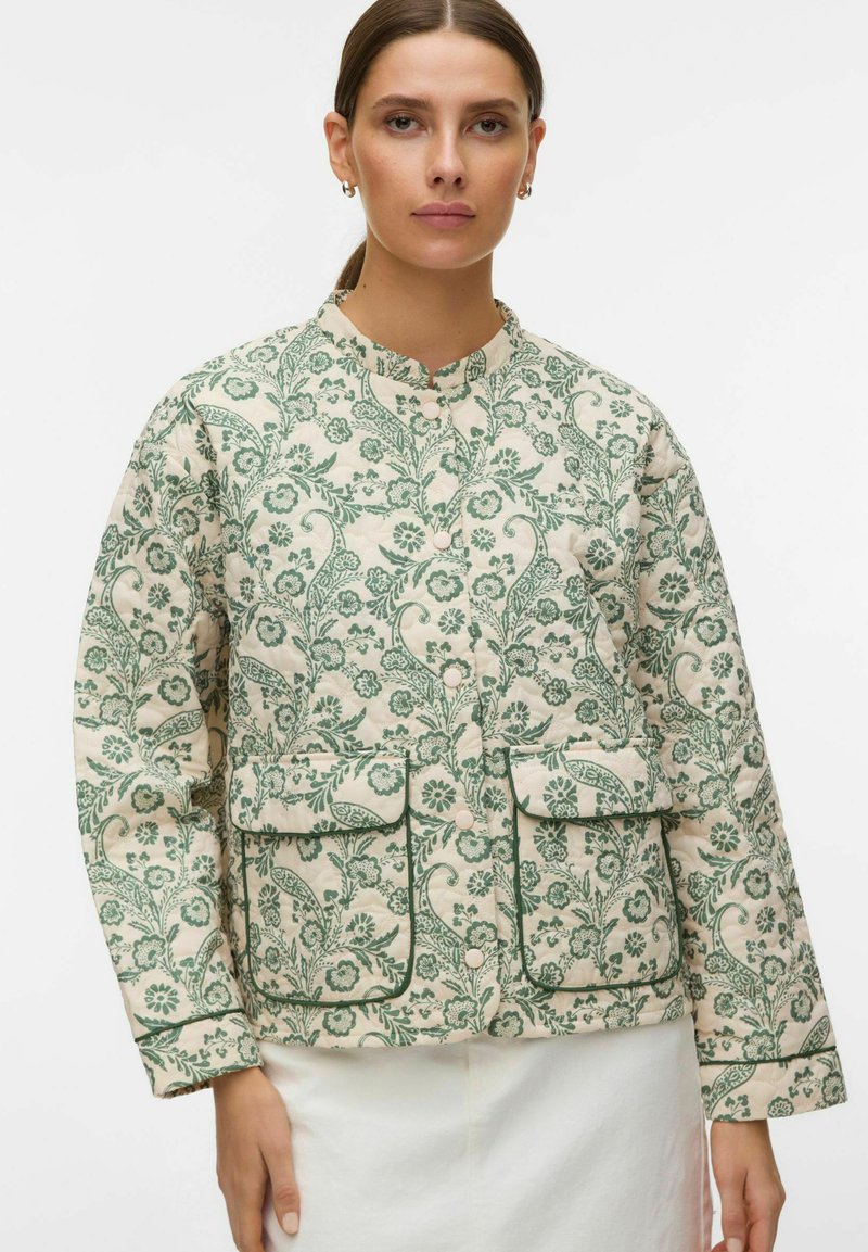 Femme portant une veste matelassée crème avec imprimé floral vert, boutons-pression, deux poches avant à rabat et col montant.
