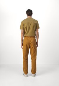 Camiseta de manga corta marrón y pantalones de pana en color mostaza. Los pantalones tienen bolsillos traseros; zapatillas blancas con suelas texturizadas completan el atuendo.