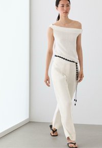Woman walking indoors wearing an off-shoulder white top, white pants with a black belt detail, and black open-toe sandals.