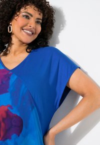 Modrá trička s barevným abstraktním designem v odstínech růžové a fialové; krátké rukávy a výstřih do V. Hladká textilie.
