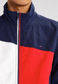 Blå, röd och vit zip-up jacka med hög krage, med färgblock design och en liten logotyp på bröstet. Slät textur.