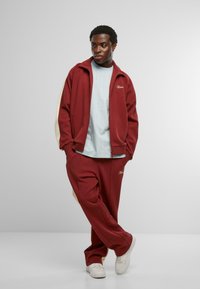 Burgundijas treniņtērps ar krēmkrāsas akcentiem, ar rāvējslēdzēju aizdaram, un saskaņoti bikses. Gaiši zila krekls ar vertikālām svītrām. Baltas kedas.