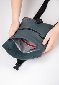 Teal waterproof bag with black zipper, tan accents, and interior red pocket. Features a single adjustable strap and top snap closure.