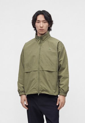 Man met zwart haar draagt een olijfgroene jas met rits en logo op de borst en zwarte broek, staand tegen een effen lichte achtergrond.