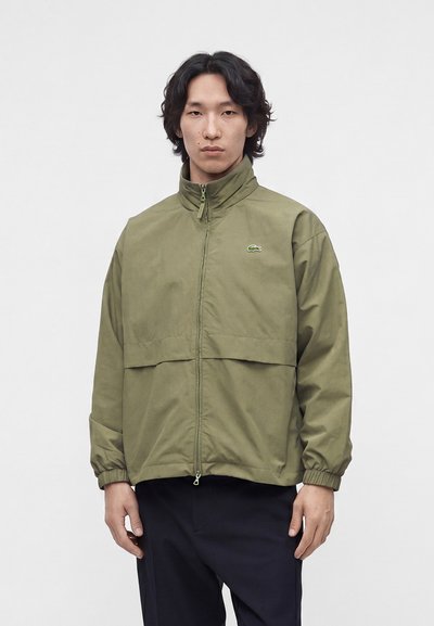 Homme aux cheveux noirs portant une veste zippée vert olive avec un logo sur la poitrine et un pantalon noir, debout devant un fond clair uni.