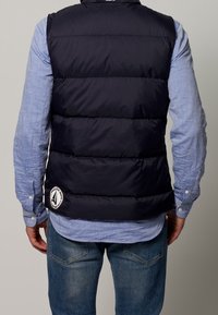 Mörkblå puffväst med horisontell sömnad, vit logotyp på nederdelen, bärs över en ljusblå skjorta med knappar, i kombination med blå jeans.