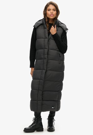 Langer, ärmelloser Steppweste in Dunkelgrau mit gestepptem Texture, Frontreißverschluss, Seitentaschen und Kapuze. Getragen über einem schwarzen Pullover und Hose.