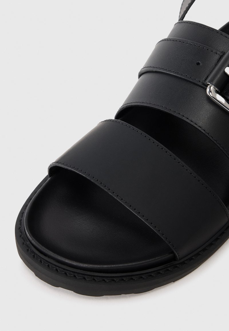 Black leather sandal with three wide straps and a flat sole, featuring a stitched edge and a metallic buckle accent.