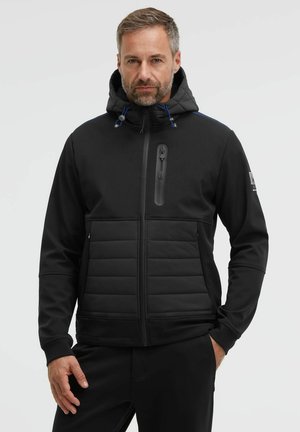 Man with gray hair and beard wearing a black hooded zip-up jacket with padded front panels and a zippered chest pocket.