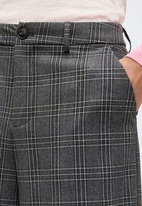 Γκρι καρό παντελόνια με μαύρο κουμπί και πλαϊνή τσέπη, φορεμένα με ανοιχτόχρωμο τοπ και χέρι που ακουμπά στην τσέπη.