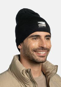Black knit beanie with a ribbed texture and a rectangular logo patch featuring a mountain scene. Worn with a light-colored jacket.