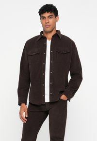 Camisa de pana marrón con dos bolsillos en el pecho, botones a presión y un cuello con solapa. Usada sobre una camiseta blanca y combinada con pantalones similares.