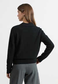 Schwarzer Pullover aus weichem, strukturiertem Stoff mit langen Ärmeln und einem Kragen, kombiniert mit grauen Hosen. Einfaches, modernes Design mit tailliertem Schnitt.