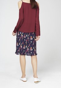 Blusa bordeaux a spalle scoperte con maniche lunghe abbinata a una gonna a pieghe blu navy con fiori, caratterizzata da un orlo arricciato e scarpe flat bianche.