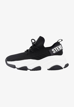 Chaussure de sport noire avec un upper en tricot texturé, semelle blanche rembourrée et une sangle avec "STEVE" en lettres majuscules à l'arrière.