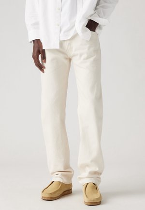 Persoon die off-white broek, lichtbeige suède mocassins en een wit overhemd met knopen draagt, met één hand in de zak.