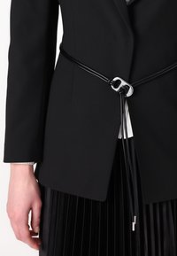 Black leather belt with a silver buckle, featuring a knotted design and hanging tassels, worn over a tailored black blazer.