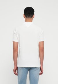 Cămașă albă din bumbac, cu mâneci scurte, guler rotund și un accent triangular în spate. Pur tată cu blugi din denim bleu deschis.