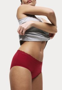 Czerwone bawełniane majtki z koronkową talią, o gładkiej fakturze i niskim stanie, w połączeniu z szarym bezrękawnikiem podnoszonym do góry.