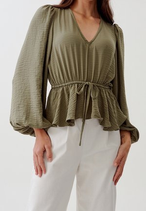 Vrouw draagt een olijfgroene blouse met lange mouwen, V-hals, trekkoord in de taille en witte broek tegen een effen achtergrond.