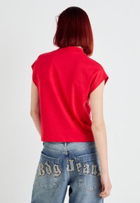 Rød kortermet croppet topp med høy hals, parret med blå denimjeans som har brodert "Big Jeans" på baklommene.